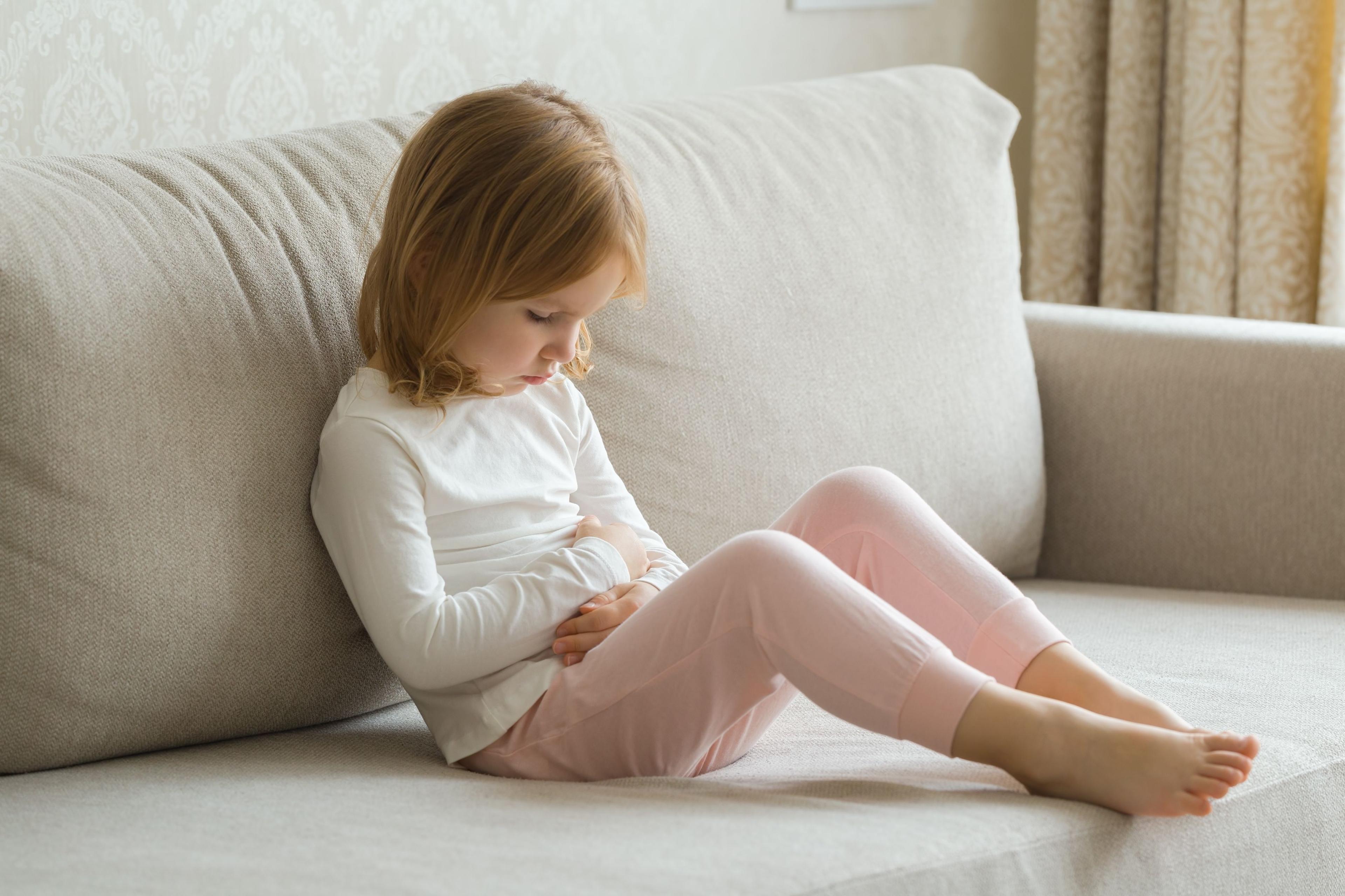En liten flicka i vit tröja sitter i en soffa och håller händerna på magen.