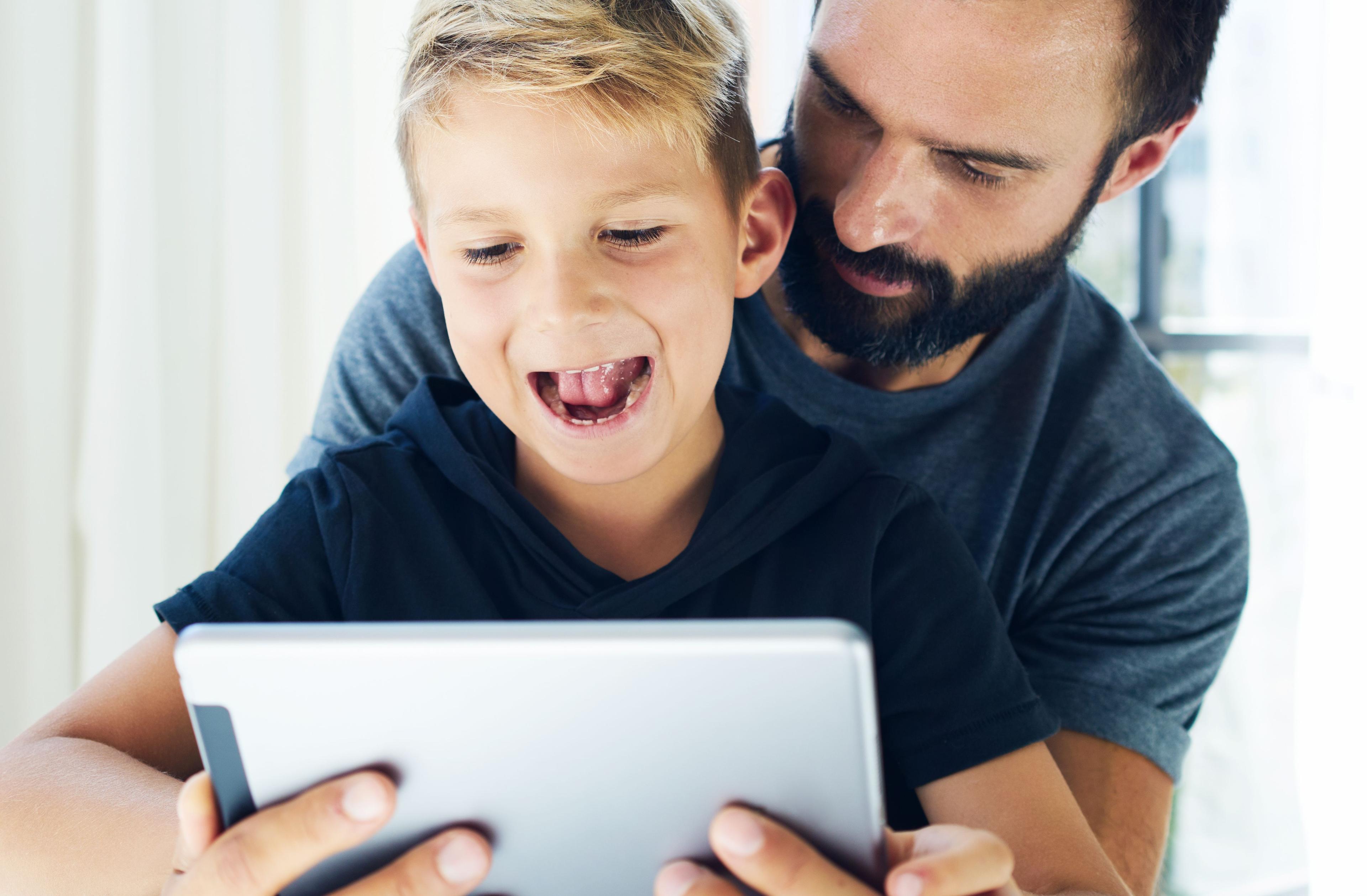 Pojke sitter i knät på sin pappa. De tittar på en Ipad.