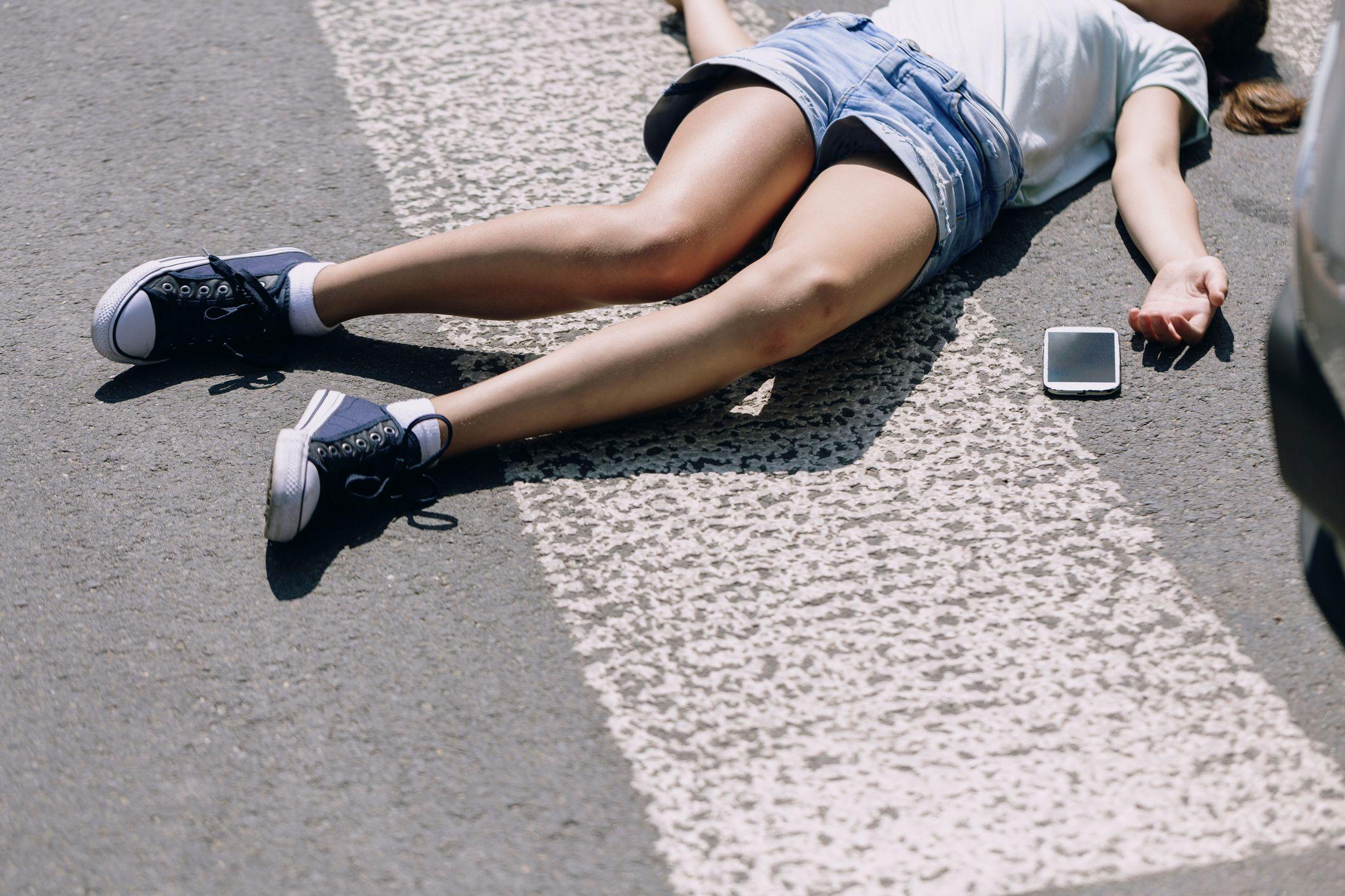 En flicka i jeansshorts och canvas-skor ligger avsvimmad på rygg på en väg. Hennes mobiltelefon ligger bredvid henne.