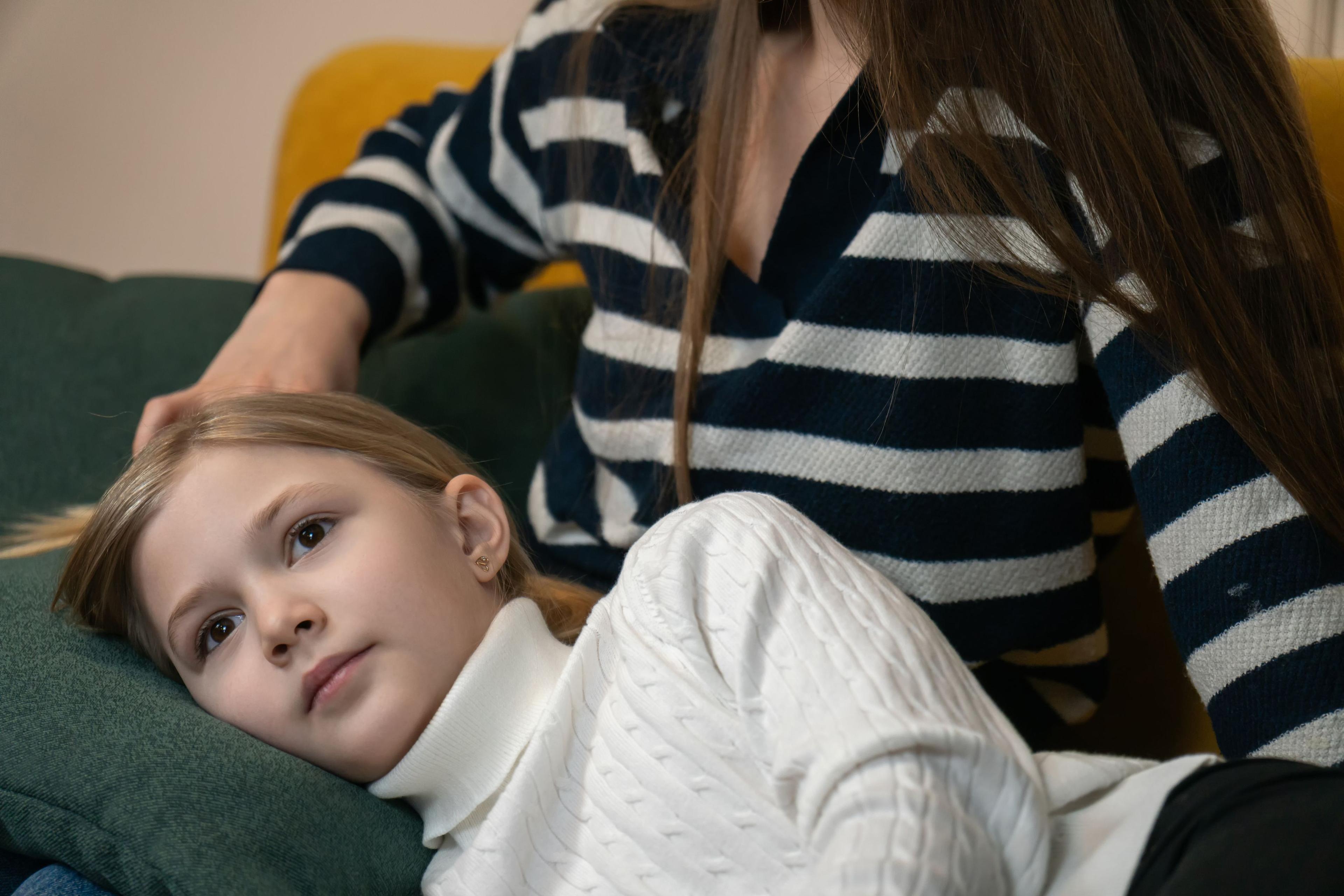 Flicka i vit tröja ligger i knät på sin mamma. Ser ut att ha ont.