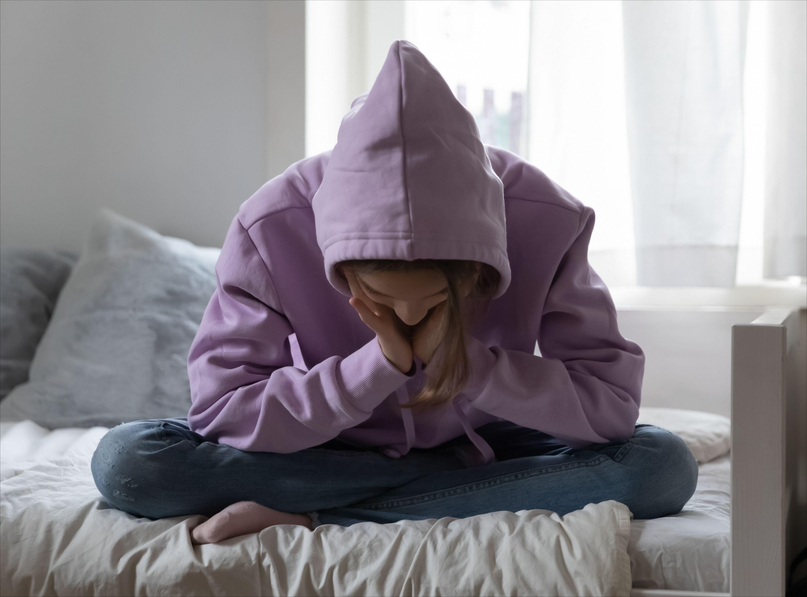 Tjej i rosa hoodie sitter i sin säng med huvudet i händerna.