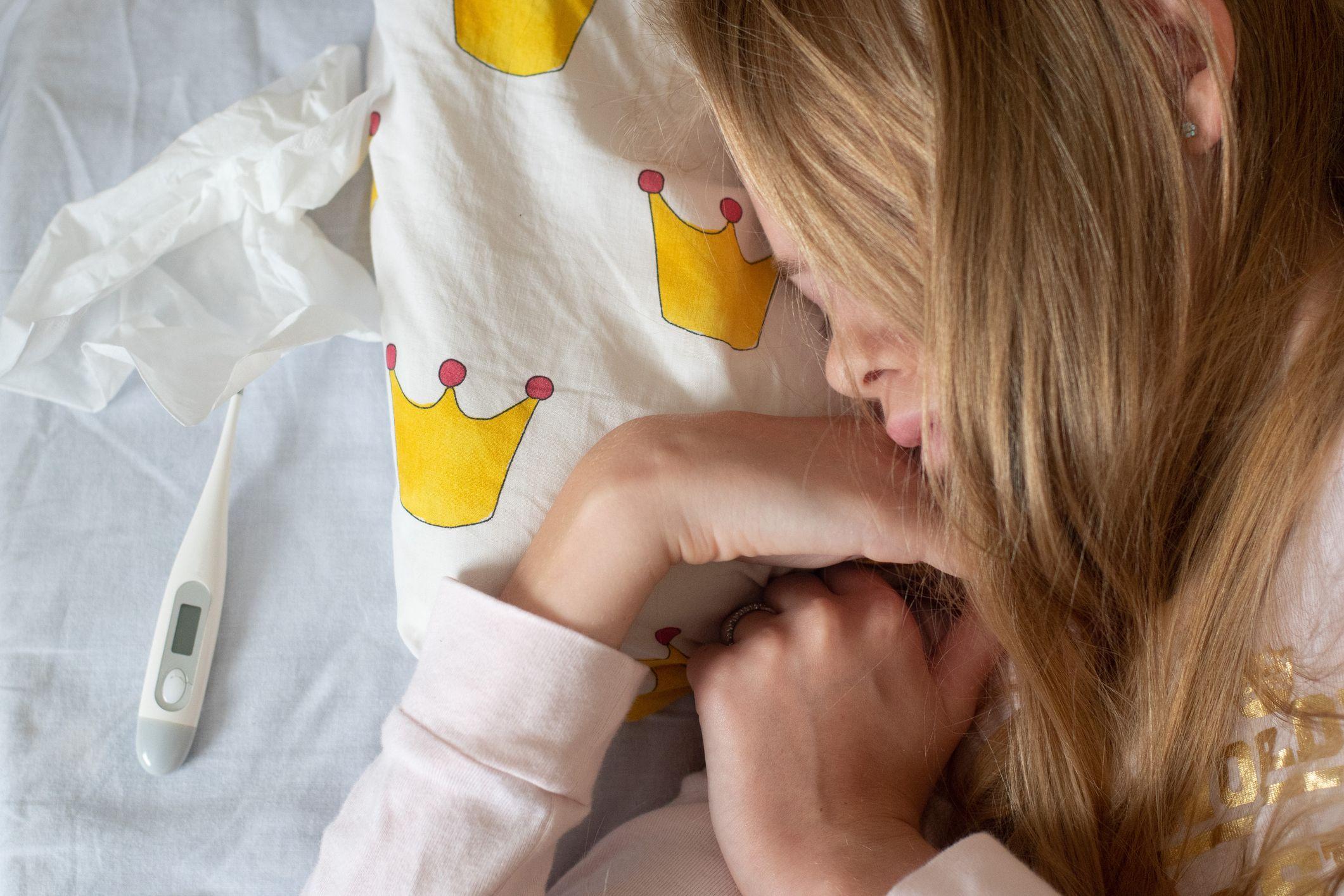 En flicka ligger på sidan i sin säng. Flickan sover. En febertermometer ligger bredvid henne på kudden.