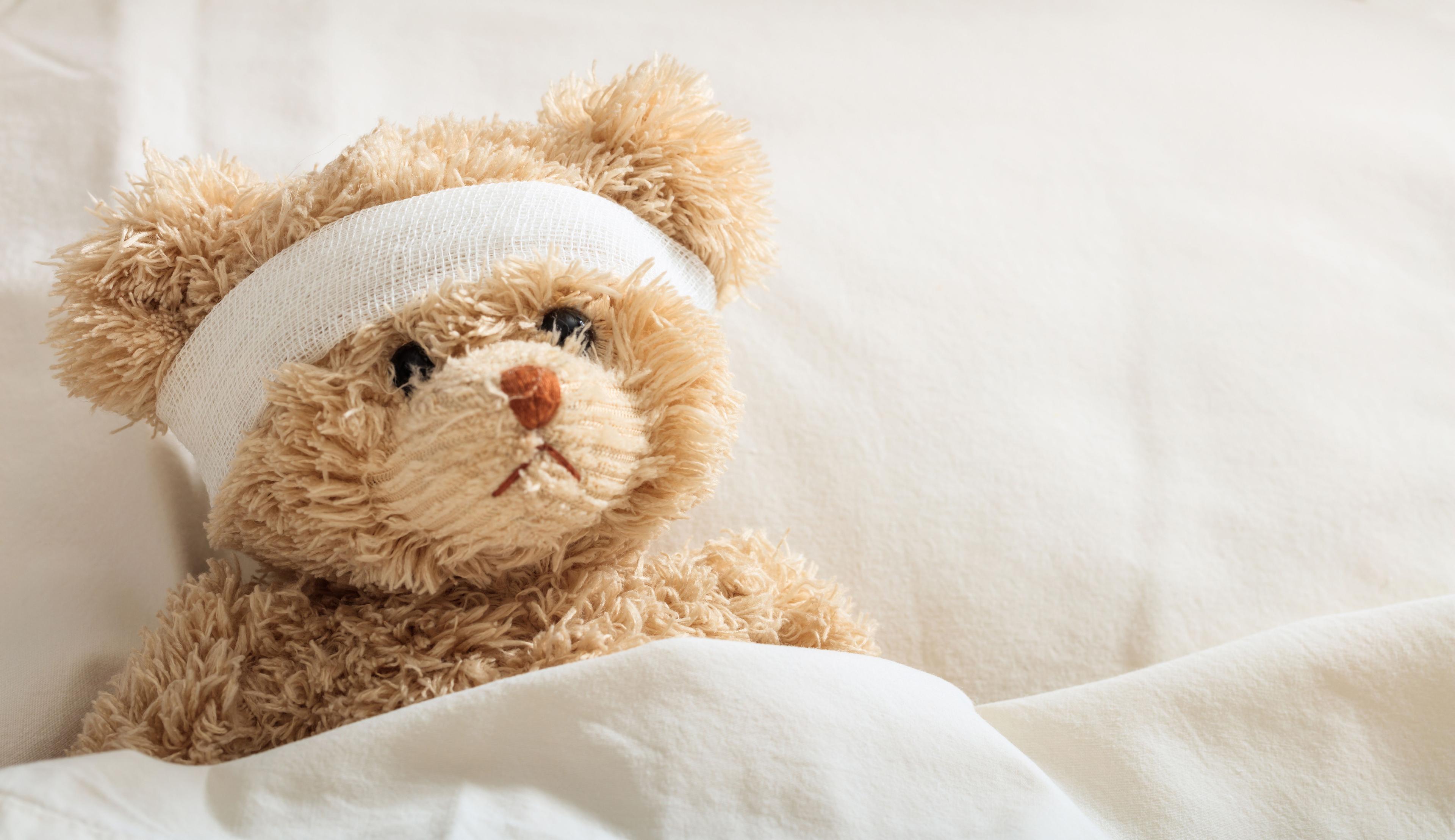 Teddy bear with a bandage wrapped around its head is lying in bed.