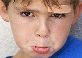 Boy in blue sweater with plaster on forehead is pouting and looking sad.