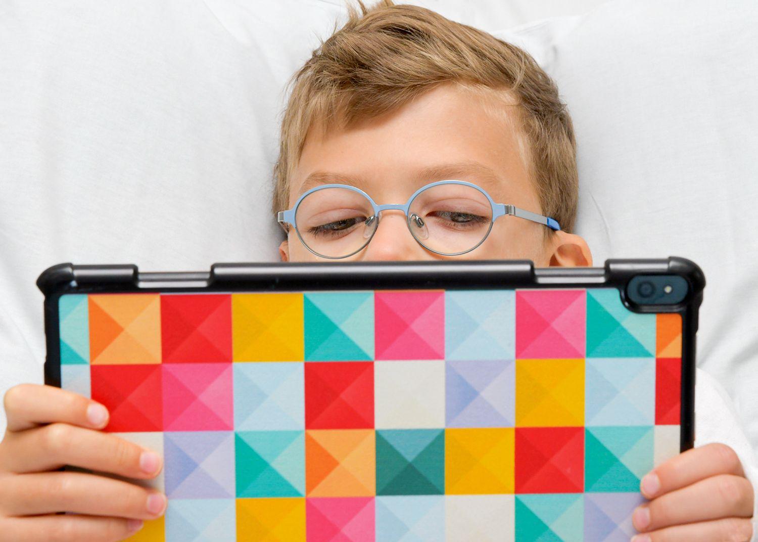 Boy with blue and round glasses is lying in bed looking at a tablet.