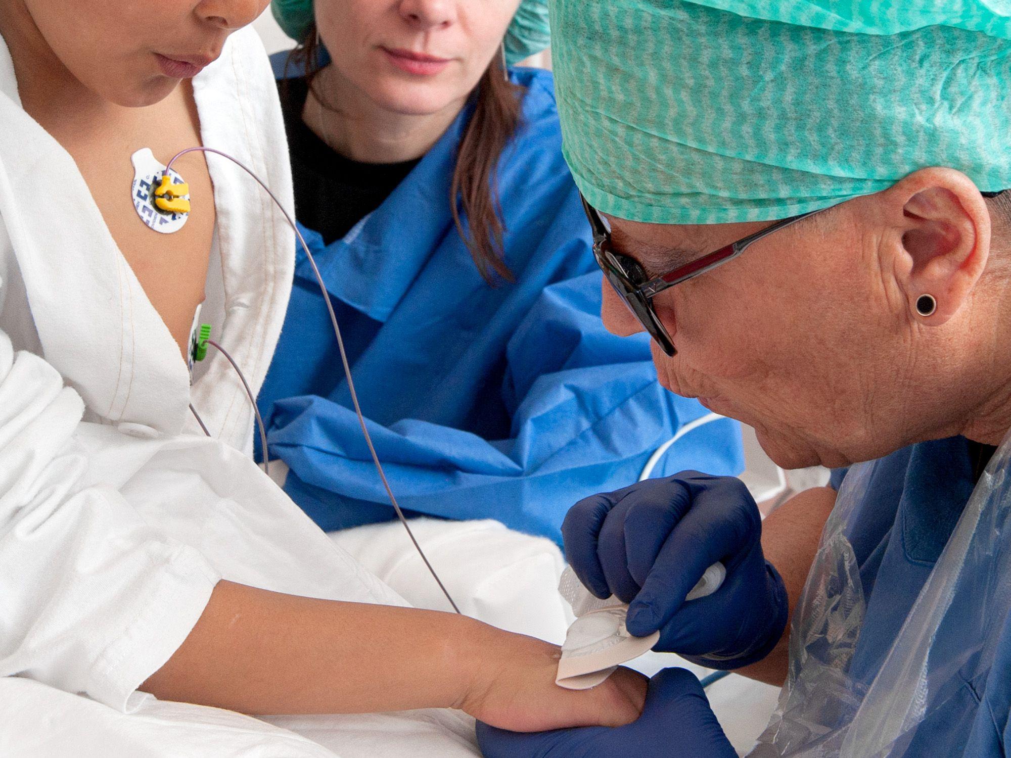 Sjuksköterska i grön mössa och blå handskar blåser på en flickas hand när hon tar bort ett bedövningsplåster.