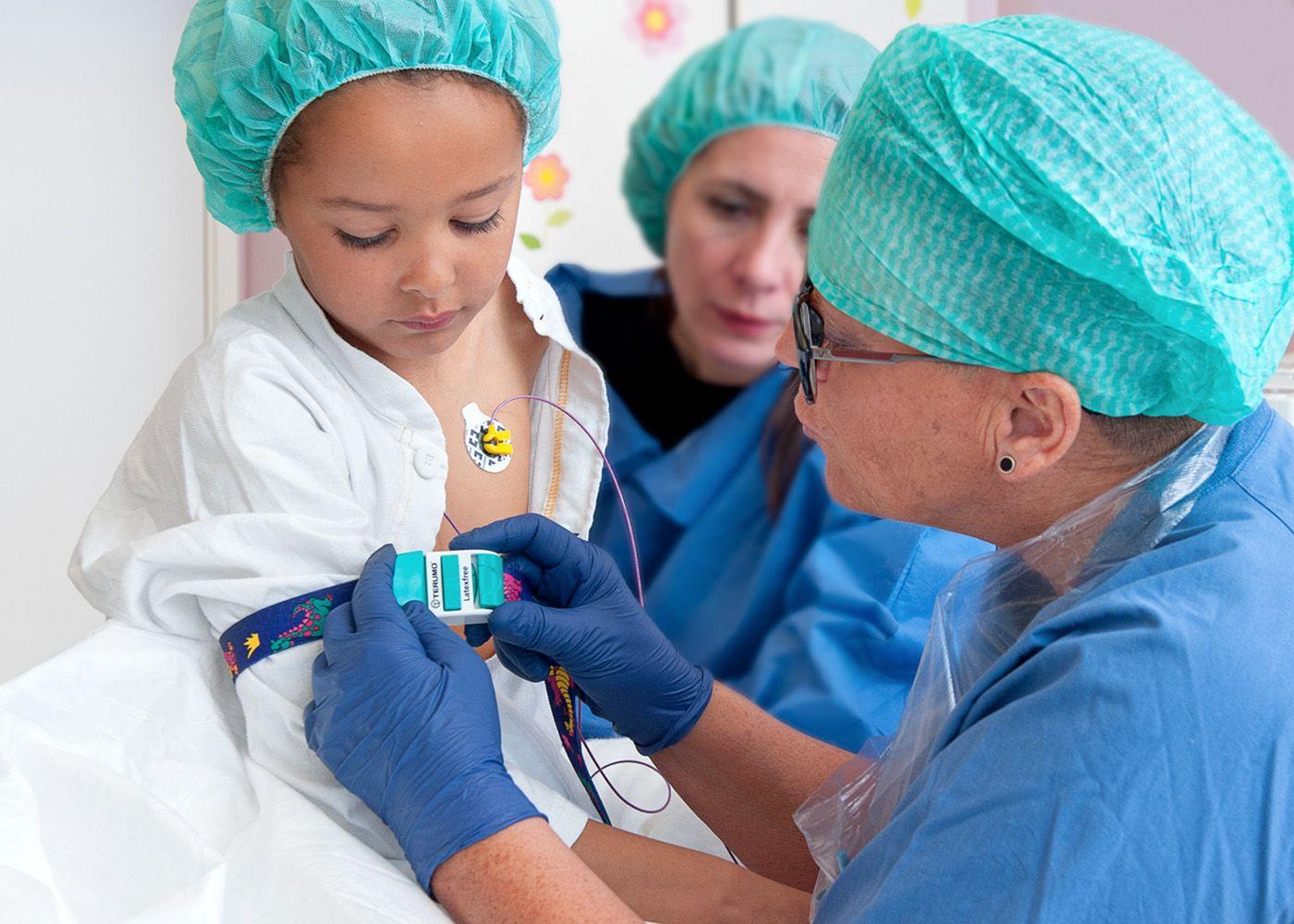 Sjuksköterska sätter fast ett stasband på armen på en flicka. Flickans mamma sitter bredvid.