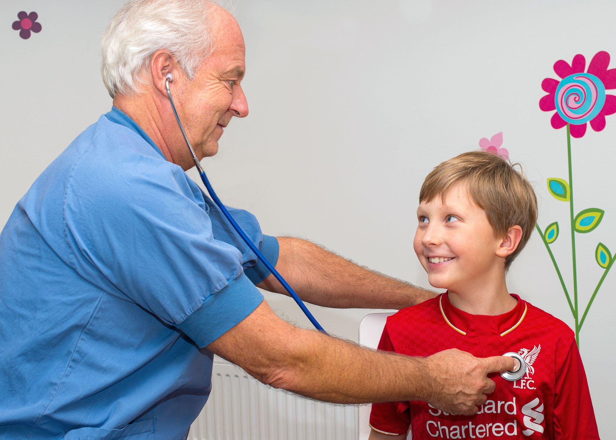 Doktor i blå operationskläder lyssnar på hjärtat med ett stetoskop på en pojke i röd tröja.