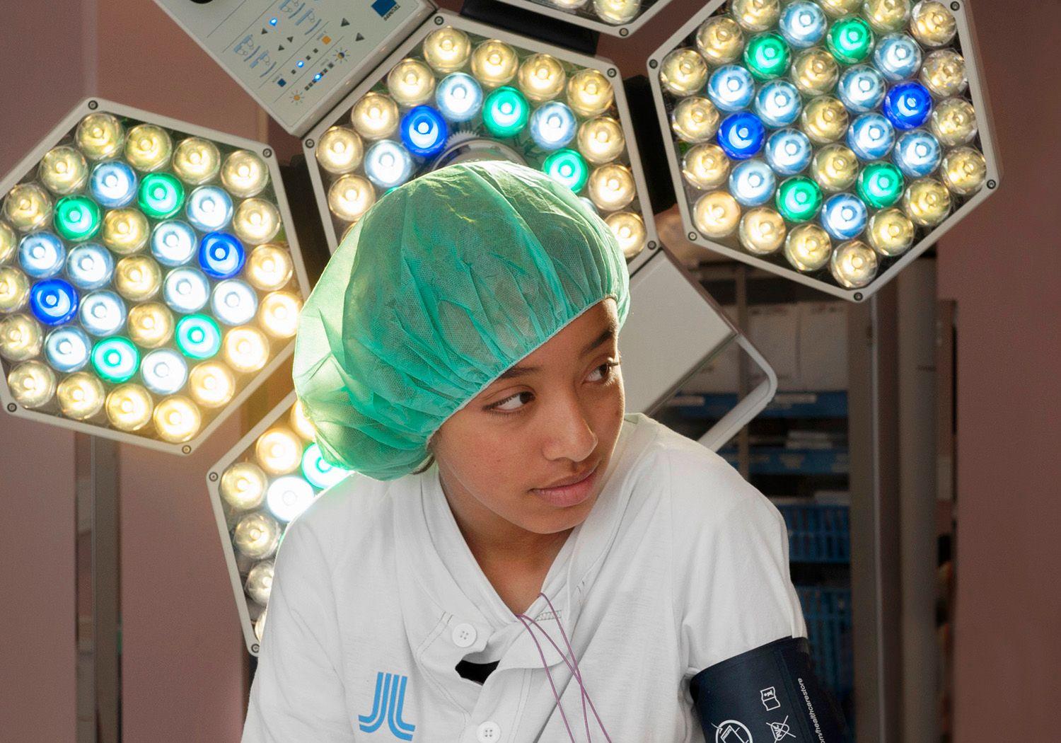 Flicka i grön operationsmössa med operationslampa i flera färger bakom sig.