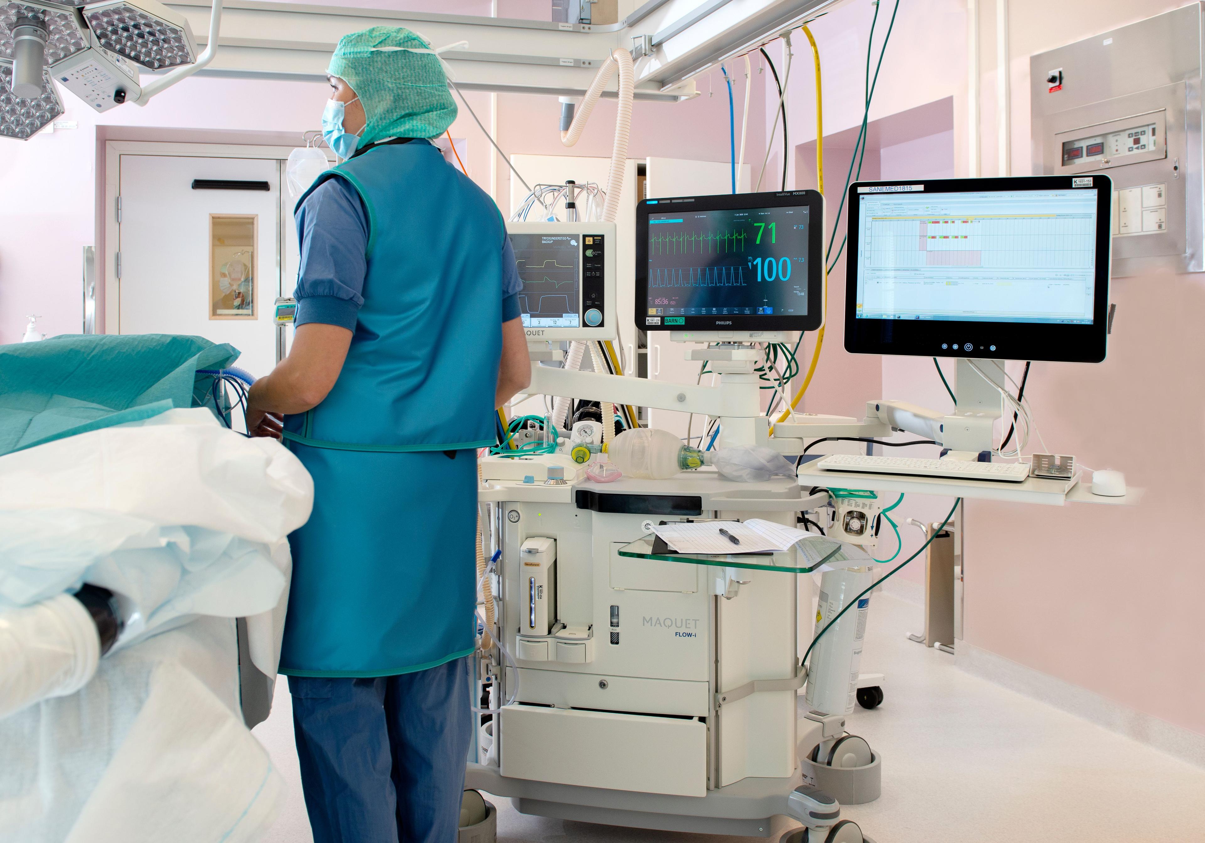 Sovmaskin på ett operationsrum. En sjuksköterska i blå kläder och grön mössa står bredvid sovmaskinen.