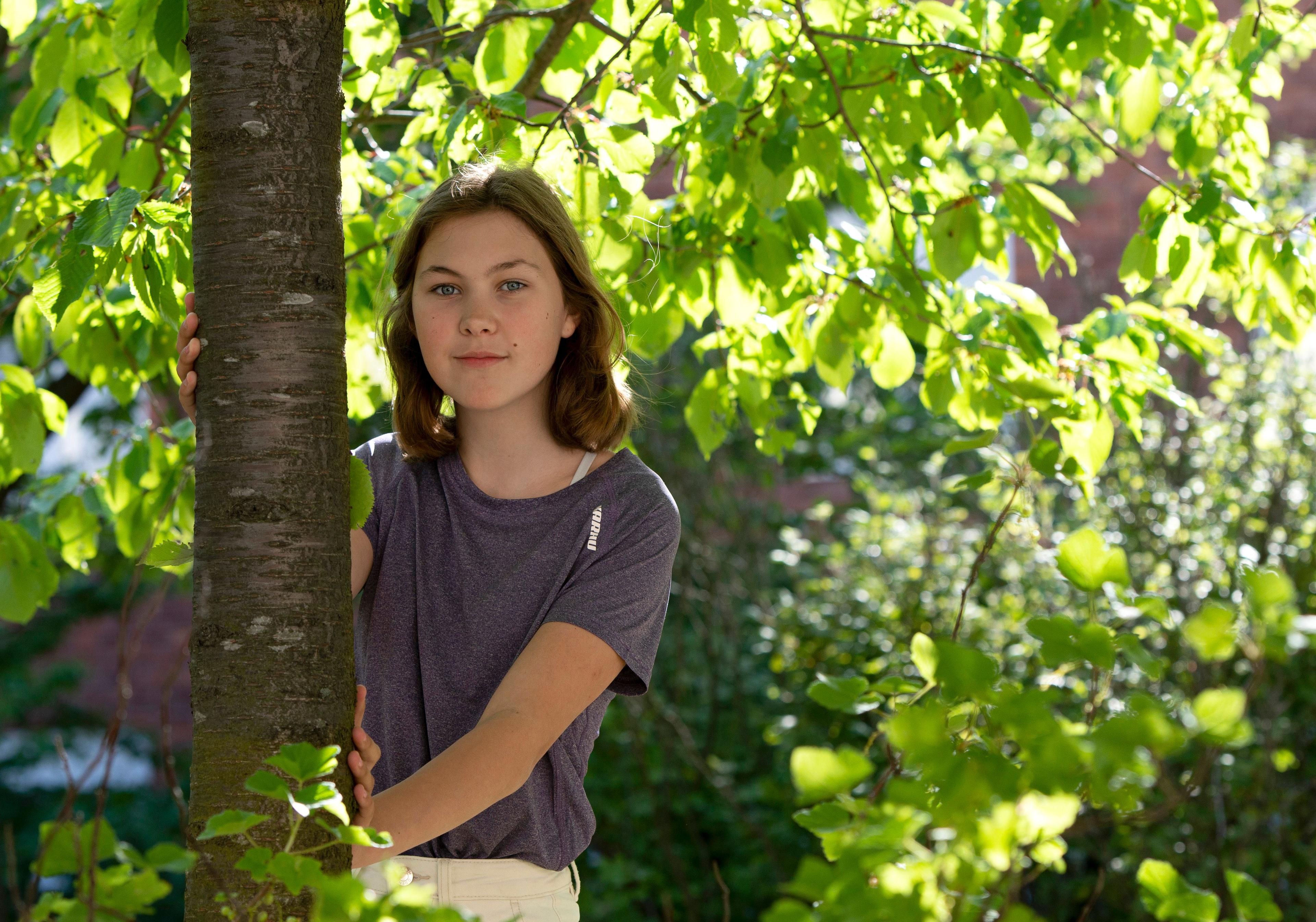 Mathilda står lutad mot ett träd, med gröna grenar och solljus i bakgrunden.