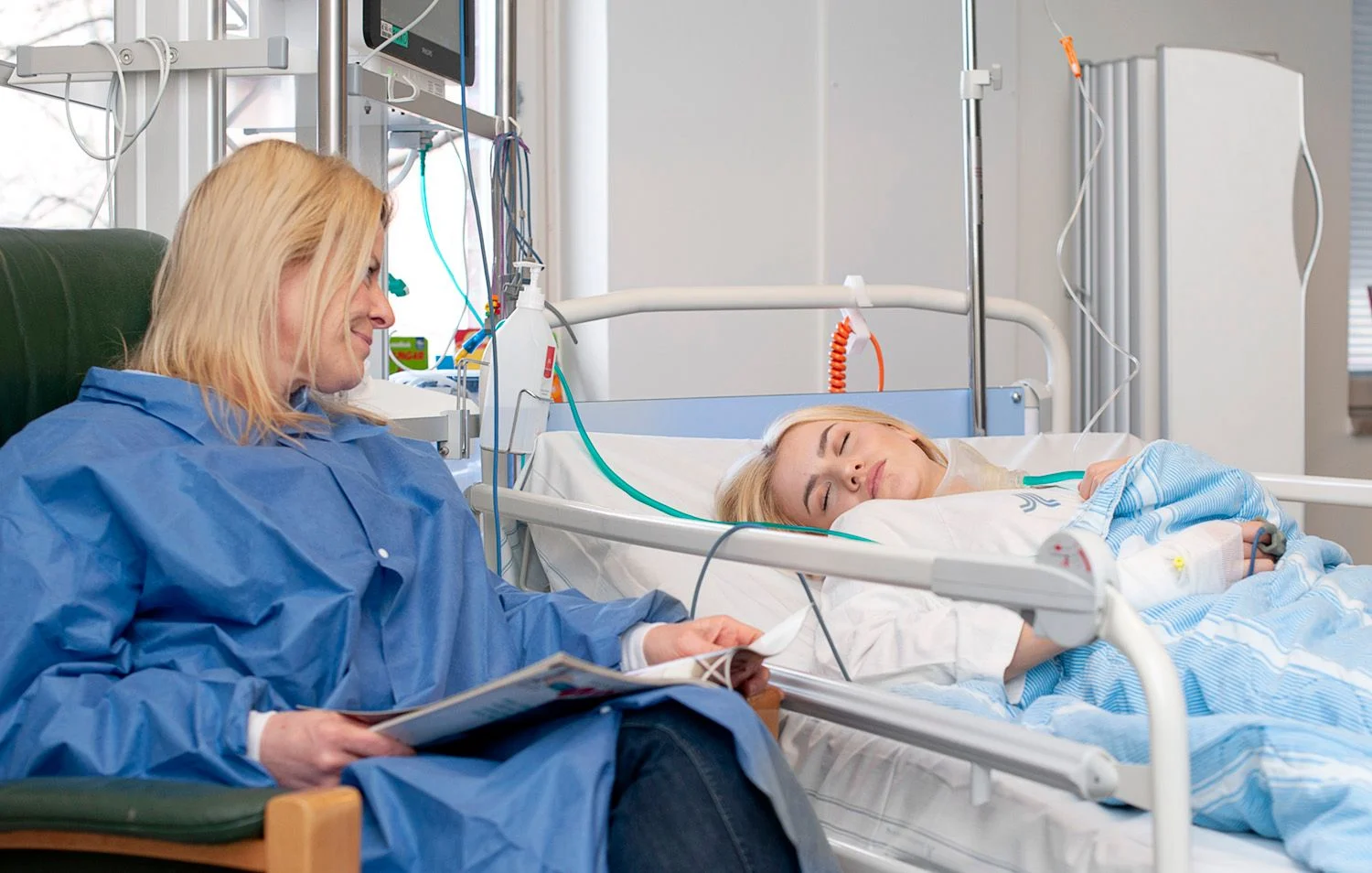 Teenage girl sleeping in her bed in the recovery ward. Her mother is sitting beside the bed.
