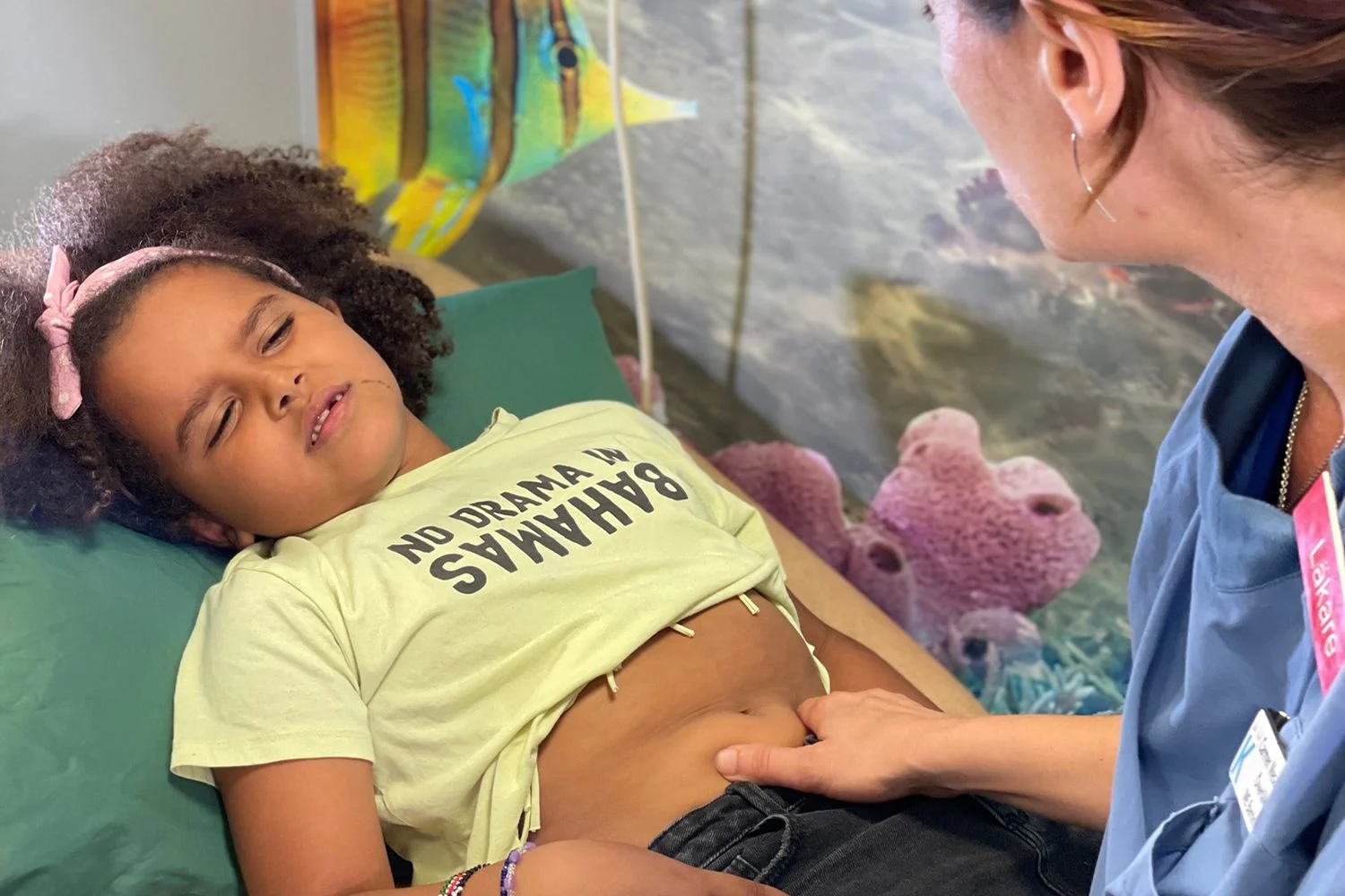 En flicka i gul t-shirt ligger på en brits på sjukhuset. Hon har ont.