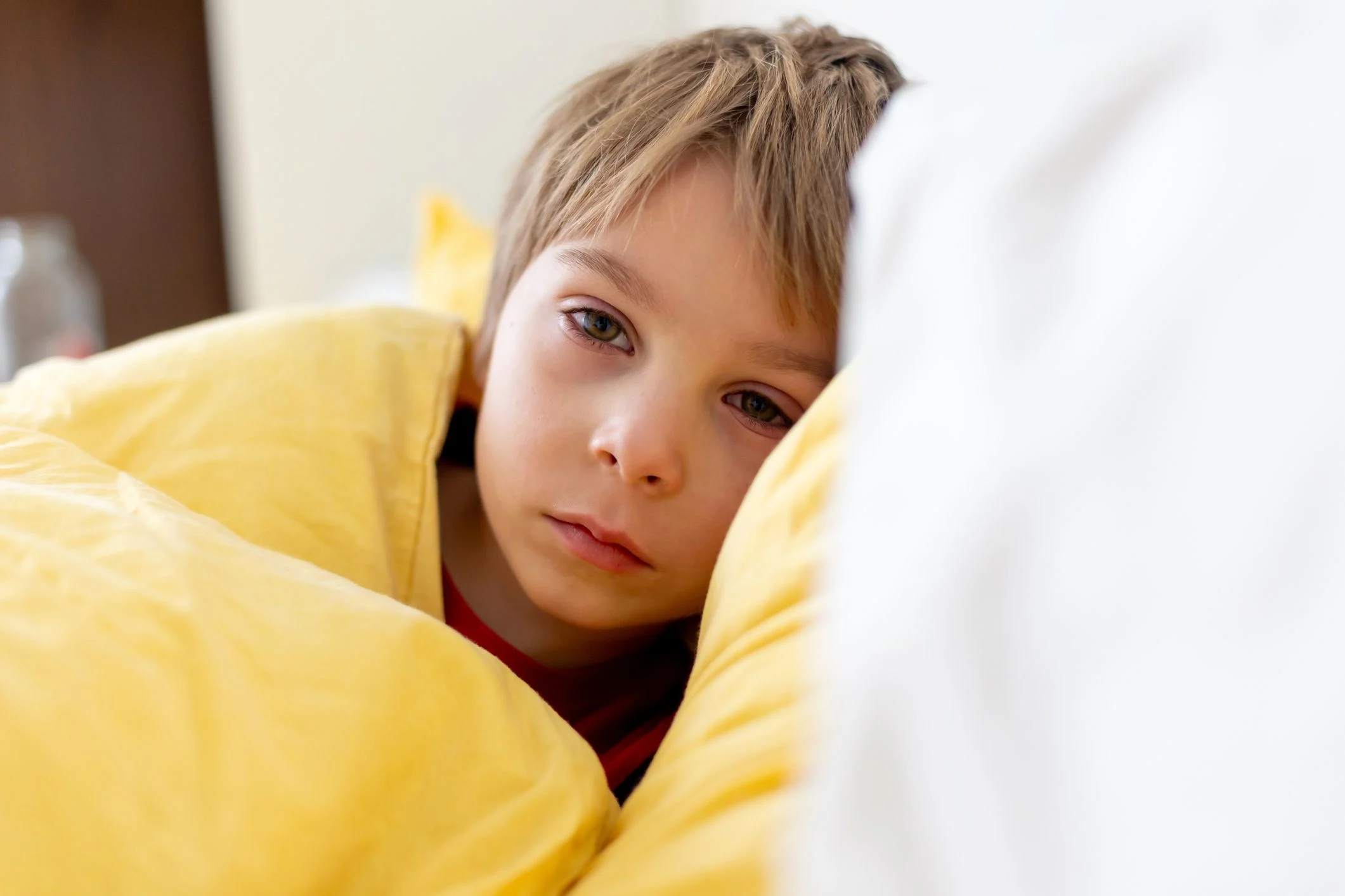 En pojke ligger på sidan under ett gult täcke i sin säng. Pojken ser trött och svag ut.