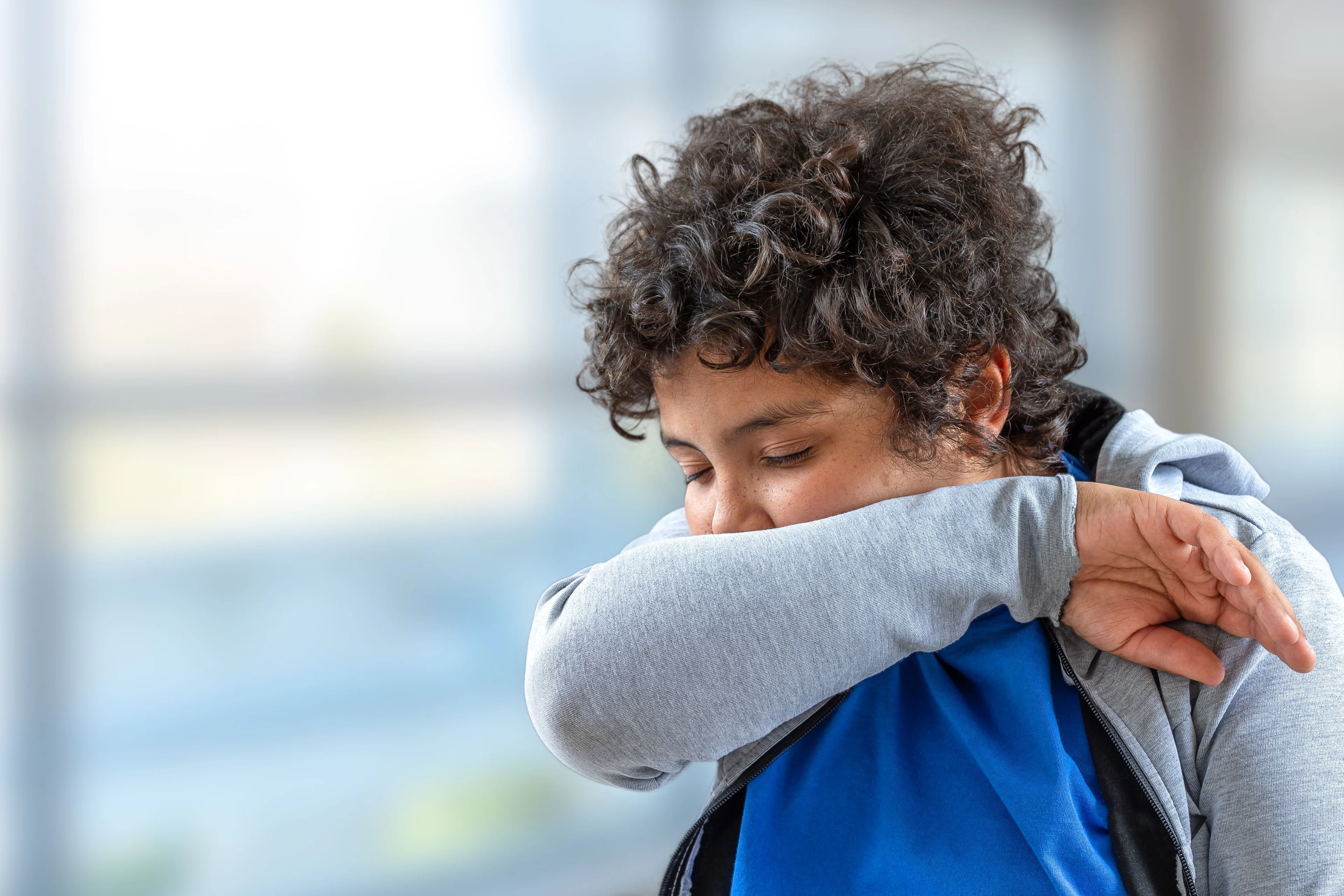 Pojke i grå tröja och mörkt lockigt hår hostar i armen.