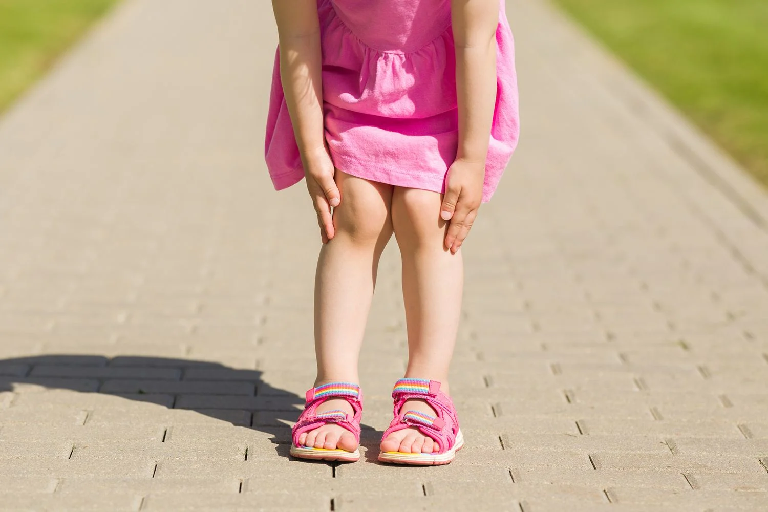 En liten flicka i rosa klänning och rosa sandaler tar sig för benen.