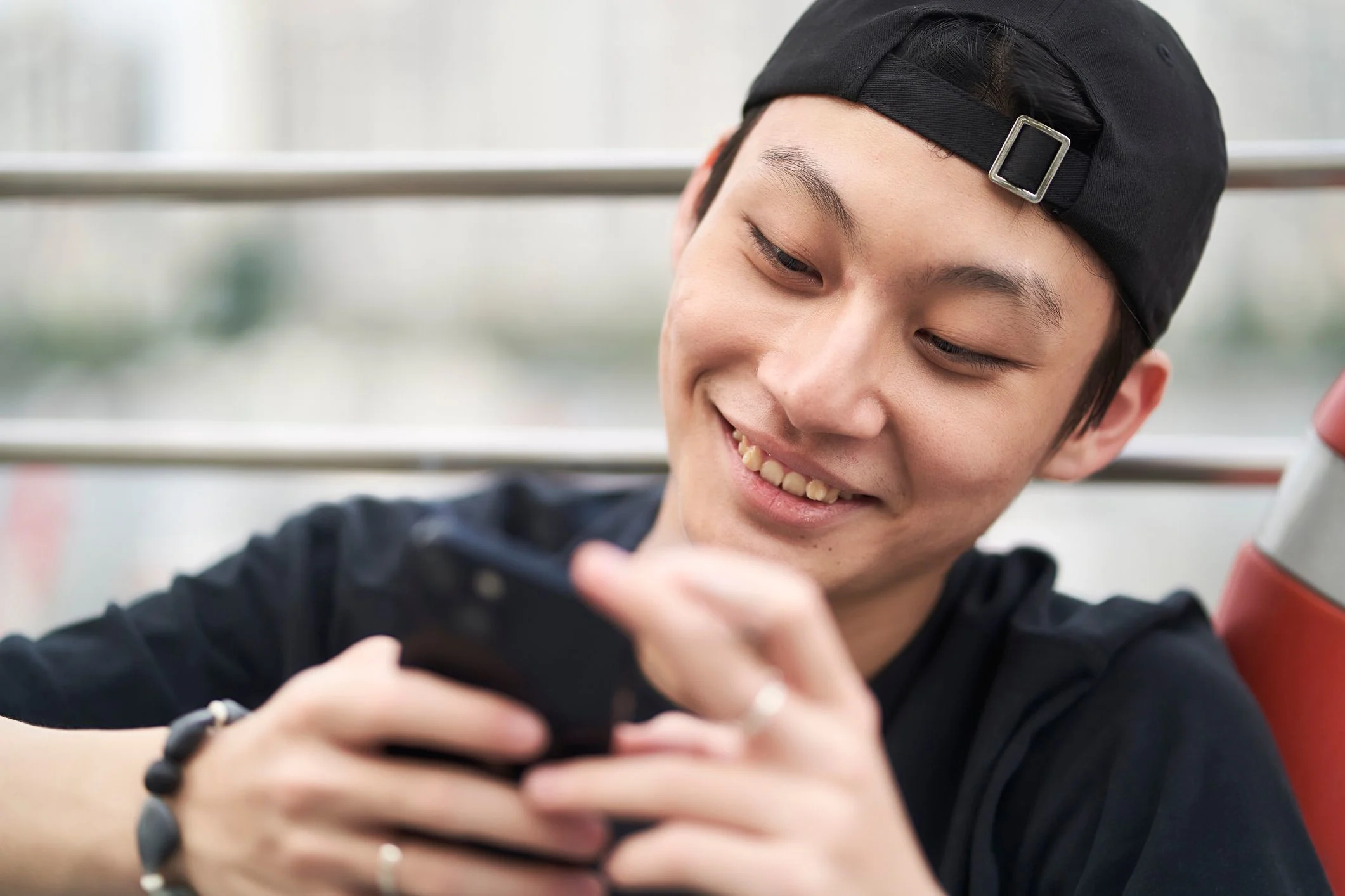 Asiatisk kille i svart kepa kollas på sin telefon. Skrattar.