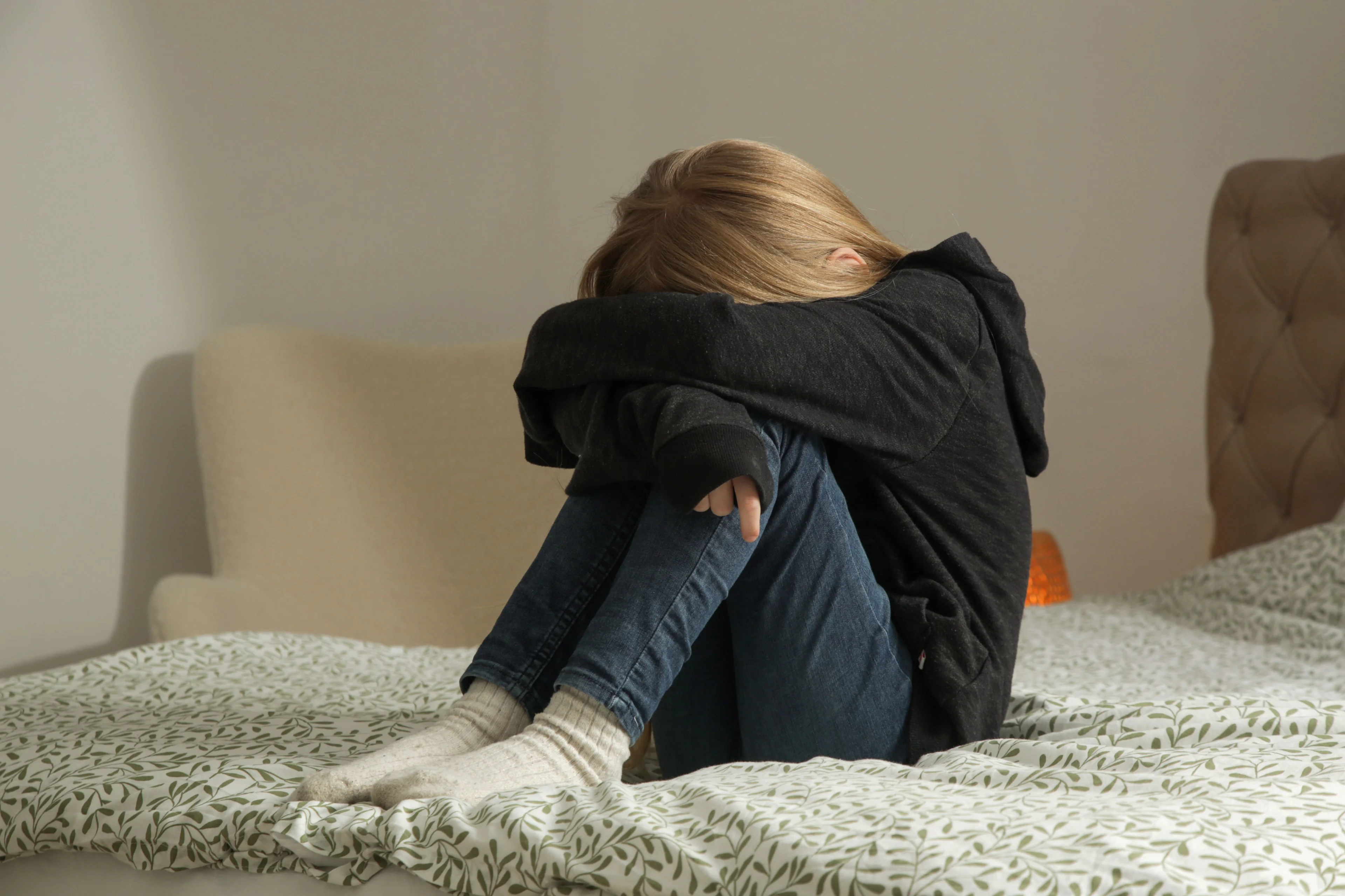 En flicka i svart hoodie sitter i sängen med uppdragna ben. Lutar sitt huvud mot benen.