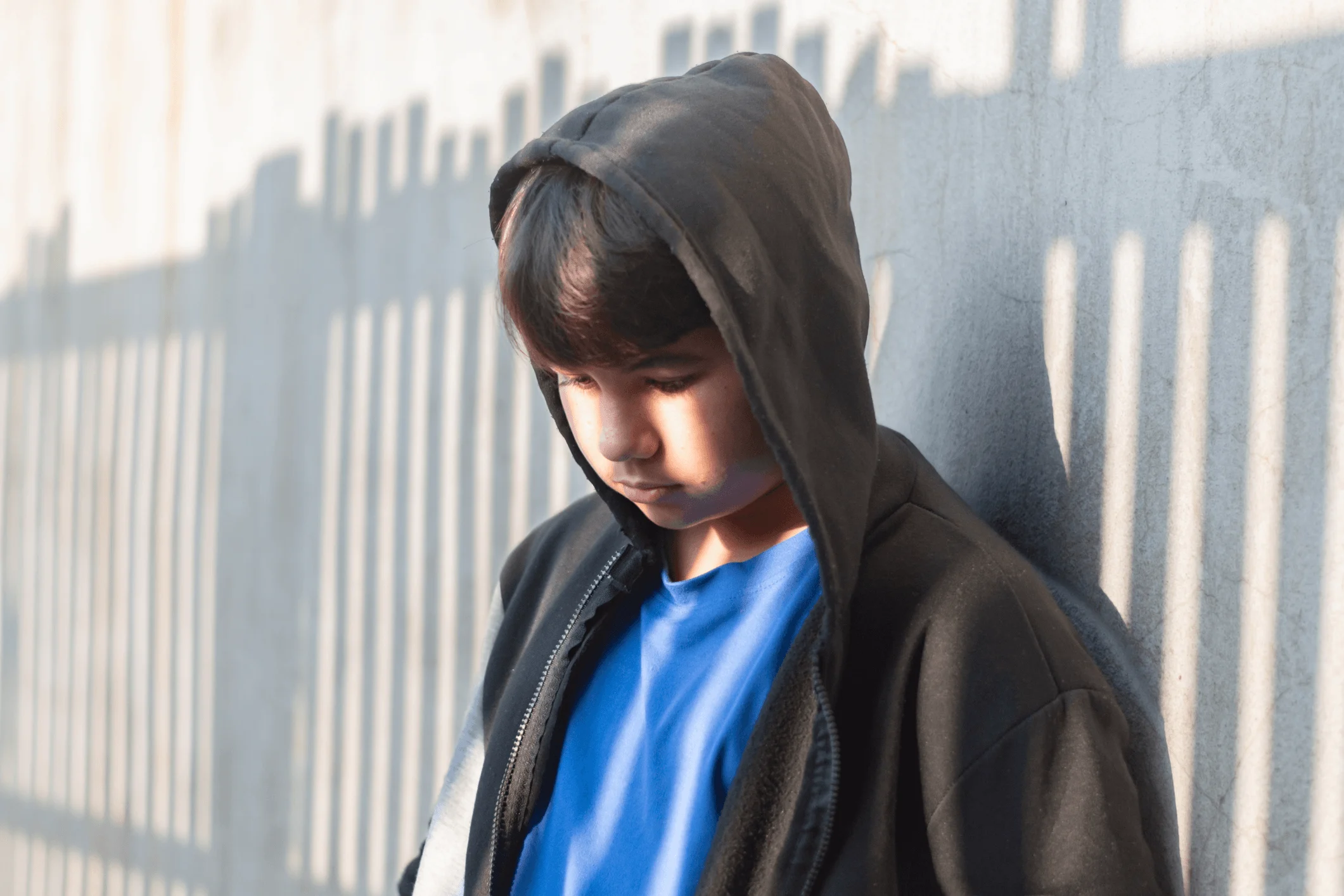Kille i svart hoodie står framför skuggan av ett staket. Tittar nedåt. Ser ledsen ut.