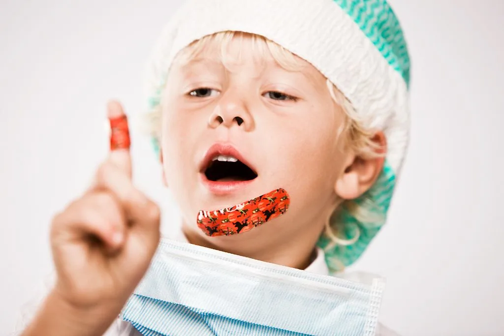 Boy wearing surgical cap and plasters on his chin and index finger. He is looking at his index finger.