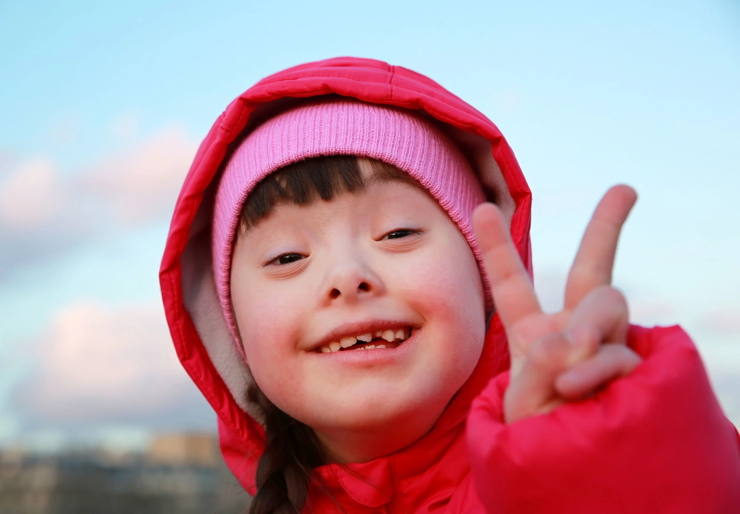 Flicka i röd täckjacka med luva gör tummen upp. Flickan har downs syndrom.