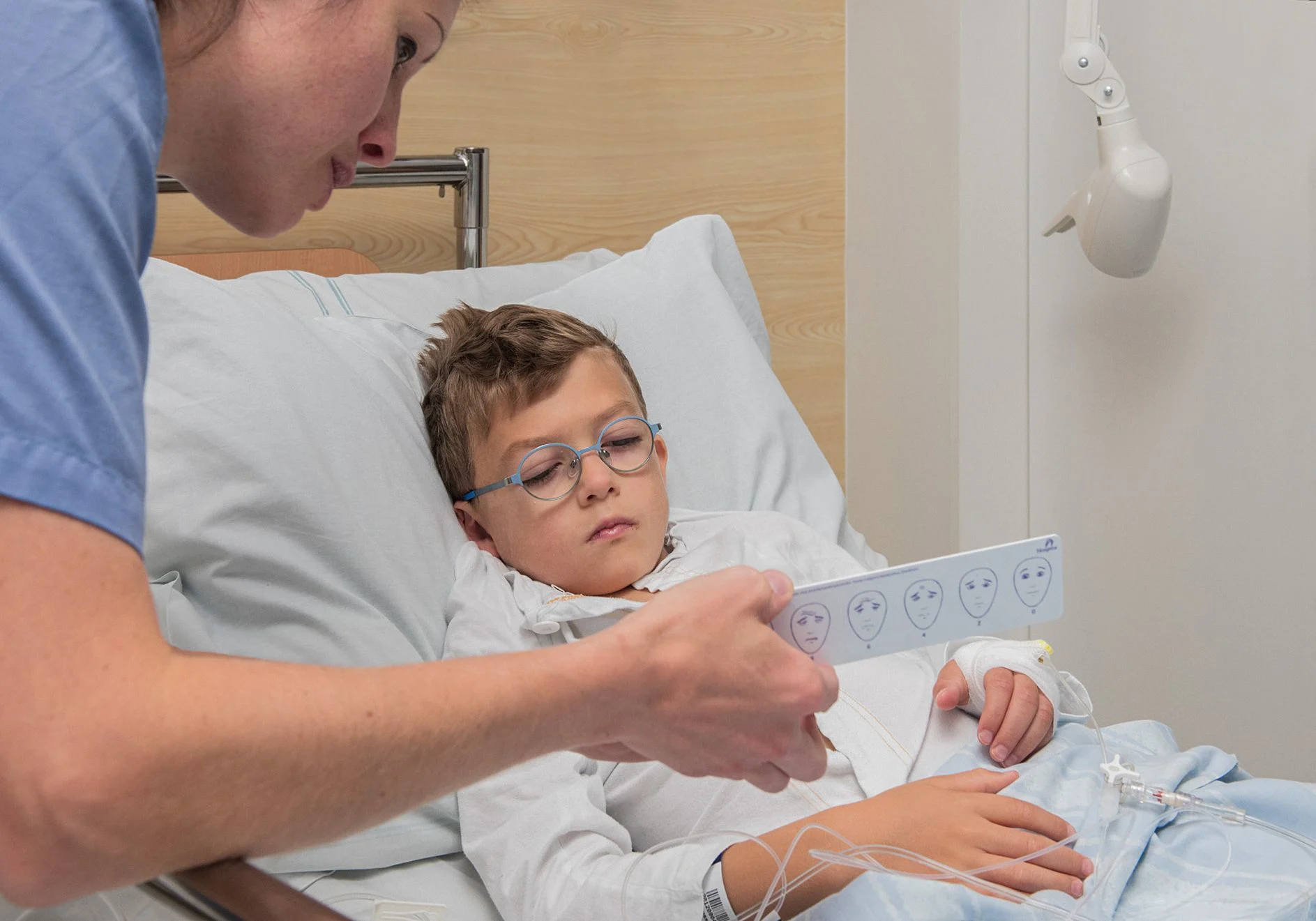 En pojke ligger i en sjukhussäng. En sjuksköterska håller en linjal med ansikten framför honom.
