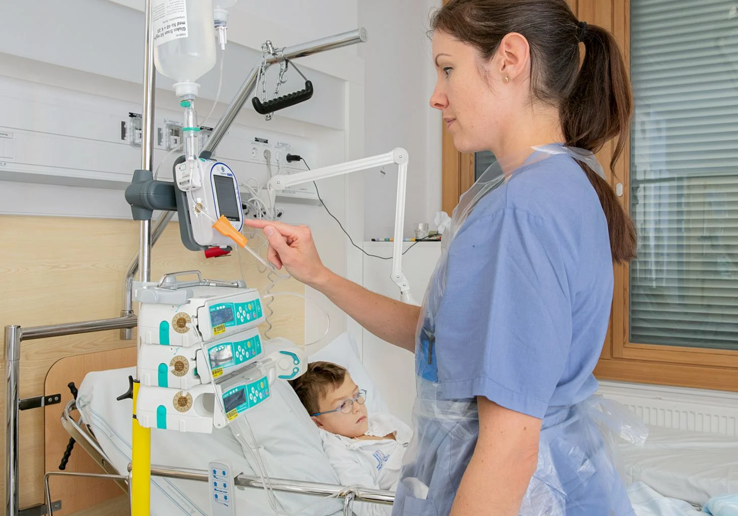 Pojke i säng med dropp kopplade till sprutpump. Sjuksköterska i blå skjorta fixar med sprutpumparna.