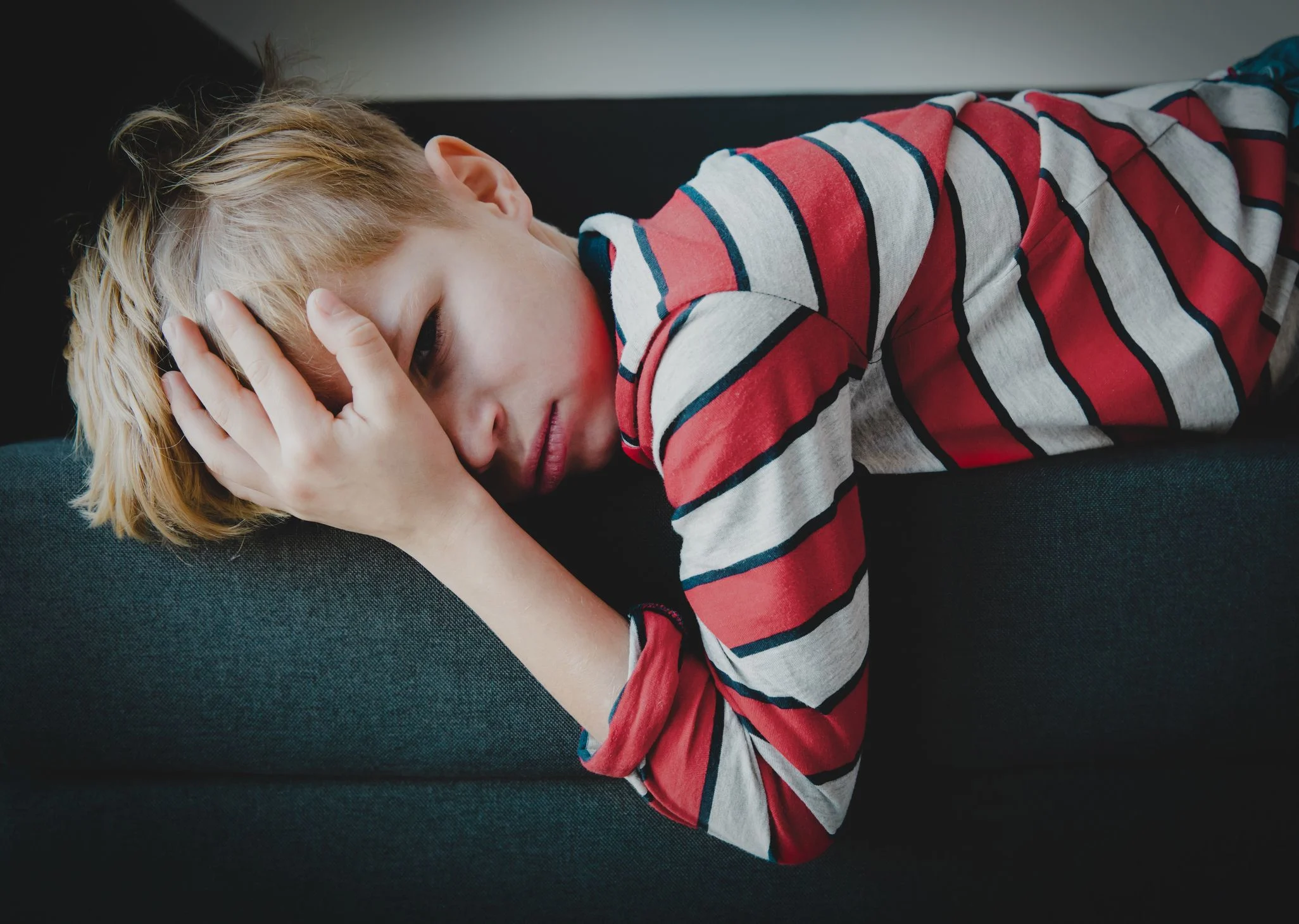 Pojke med ljust hår och röd- och vitrandig tröja ligger på magen i en soffa. Han ser trött och sjuk ut och tar sig med handen över huvudet.