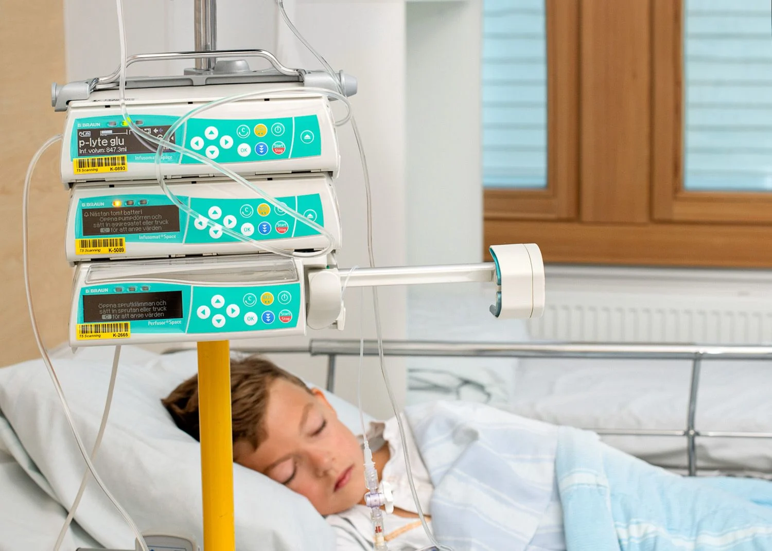 En pojke liger i en sjukhussäng. Gröna medicinpumpar i förgrunden.