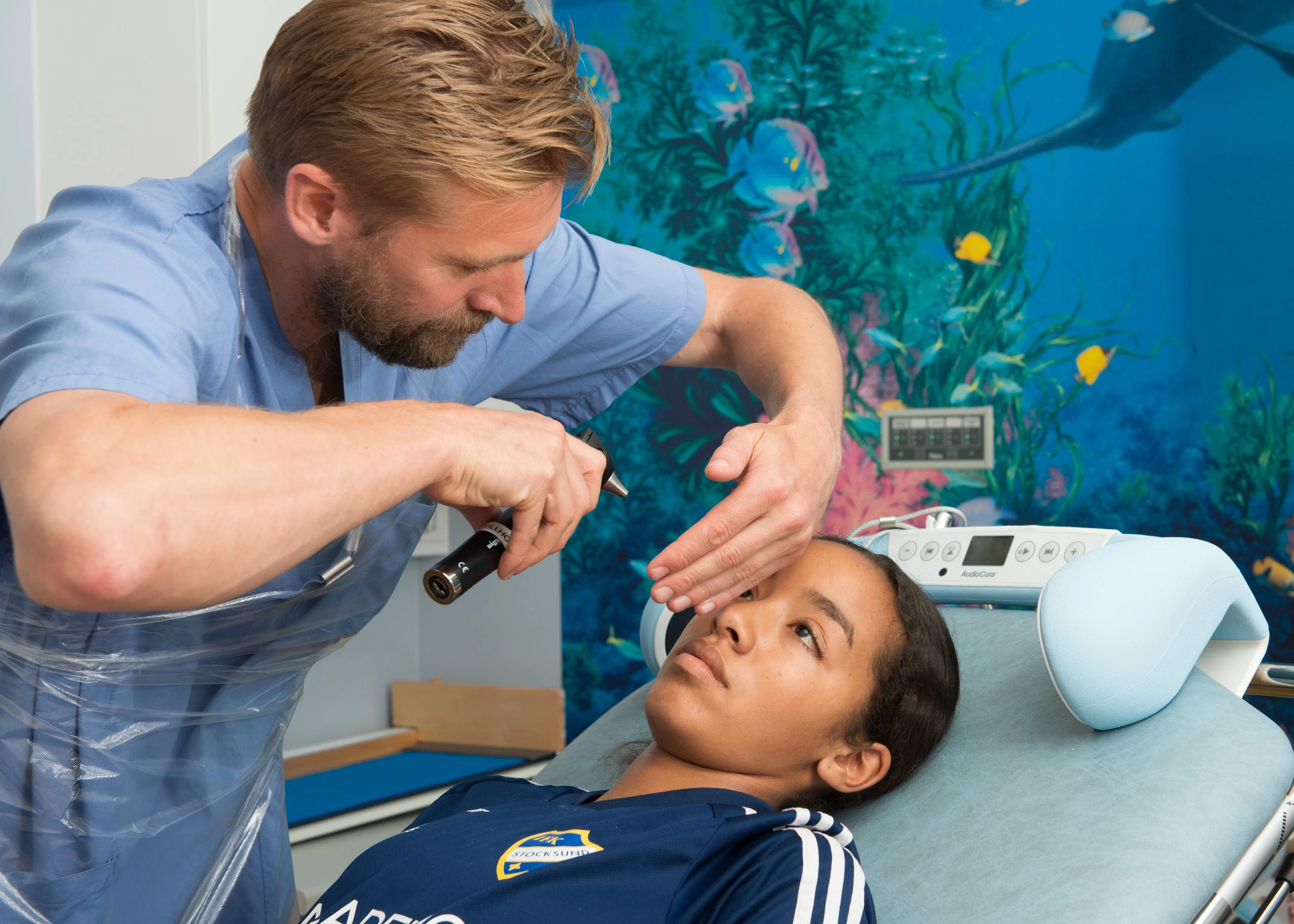 En tjej i fotbolloskläder ligger på en brits. Har fått ett slag i huvudet. En doktor undersöker hennes huvud och ögon med en lampa.