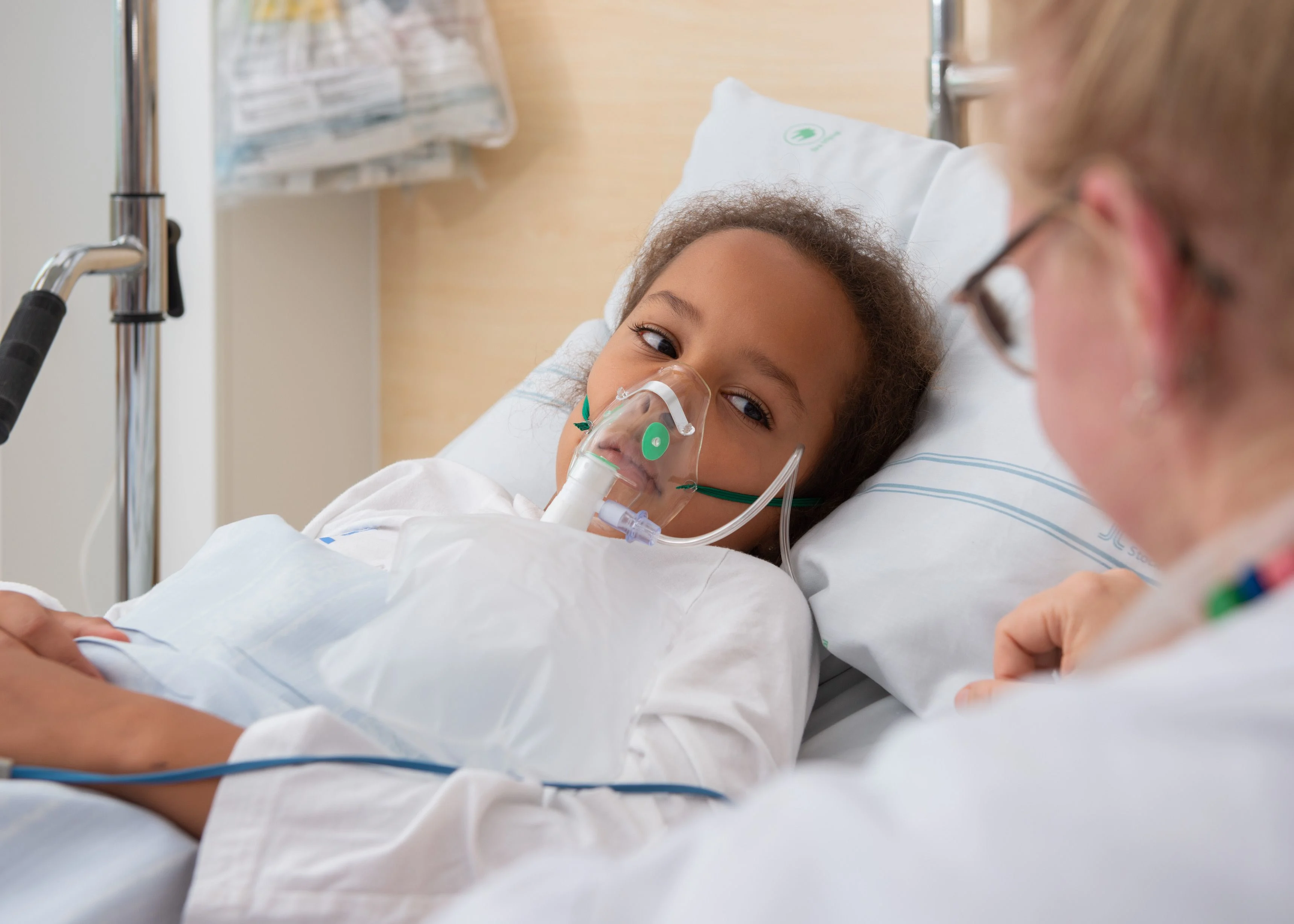 En flicka ligger i en sjukhussäng. Hon andas syre genom en tratt som hon har över munnen och näsan.