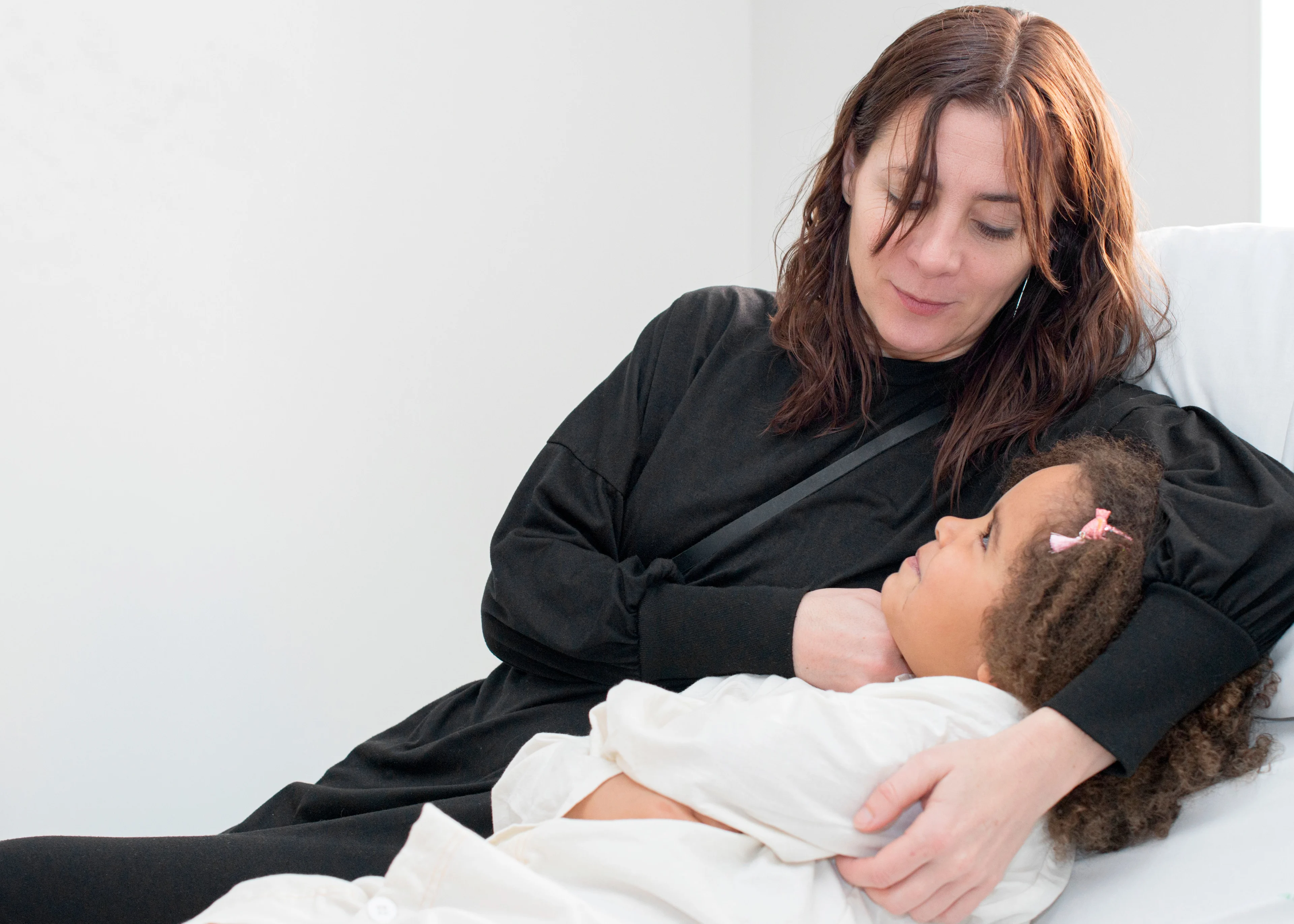 Flicka med mörkt, lockigt hår ligger i en säng. Mamma sitter bredvid.