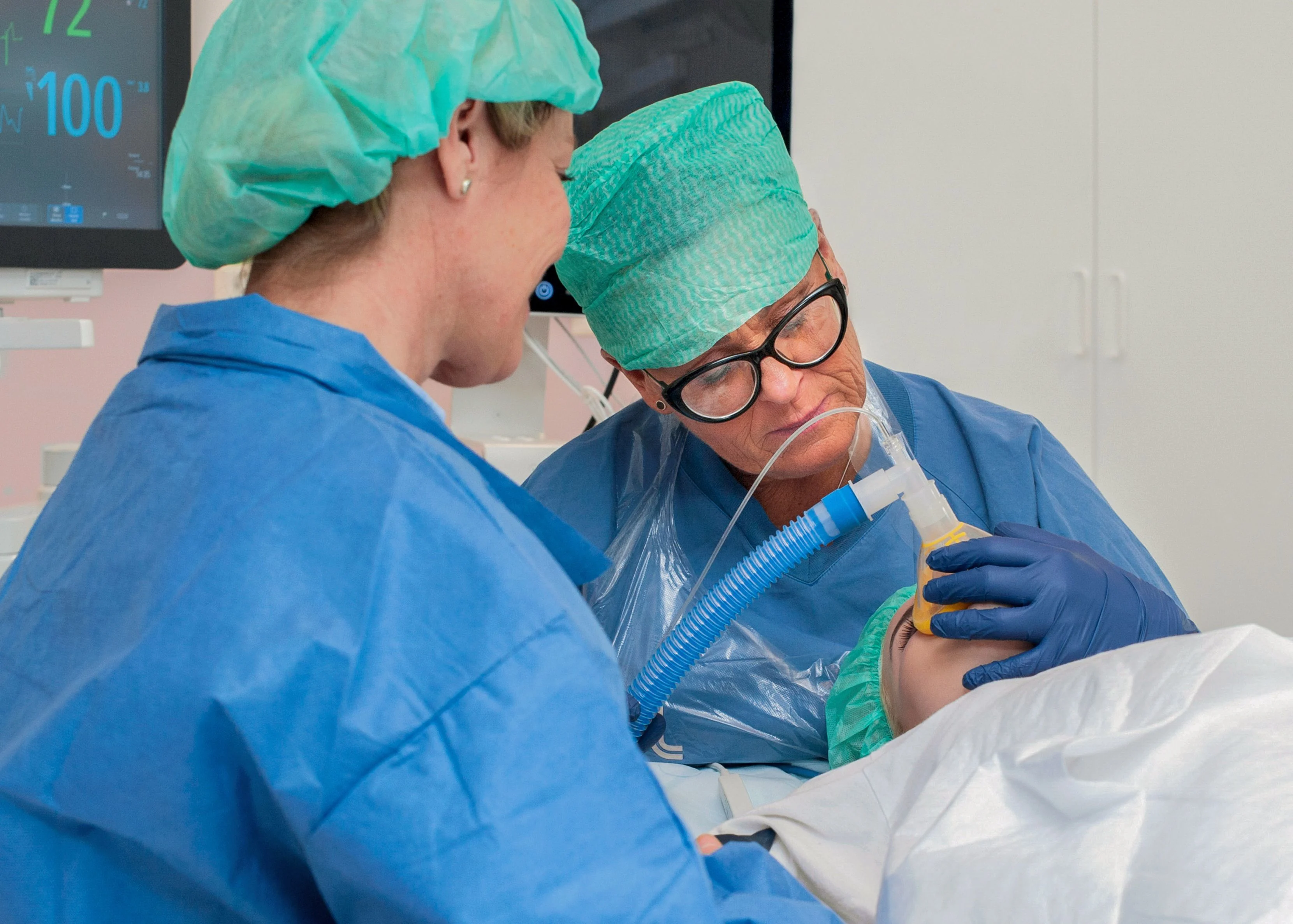 Ett barn ligger på ett operationsbord, andas i en syrgasmask. Flickans mamma sitter bredvid.